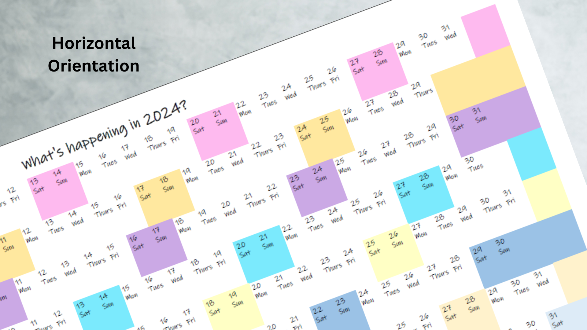 Horizontal orientation 2024 wall calendar in pastel rainbow colours shaded for all weekend dates. Framed 2024 wall calendar presented diagonally on a grey background.